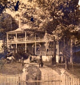 Sepia toned photograph of a two-story brick mansion with double front porches. Several people stand on the porches and the curving gravel drive in front of the house. In the close foreground is a white picket fence. The photograph was taken in 1875.